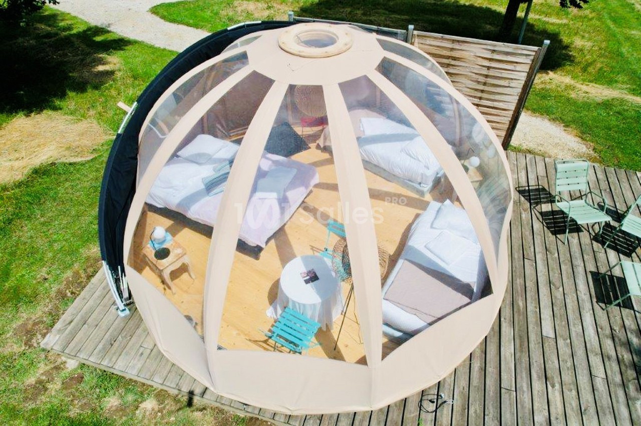 Vue aérienne d'une bulle transparente avec deux lits, une table et des chaises sur une terrasse en bois entourée de verdure.