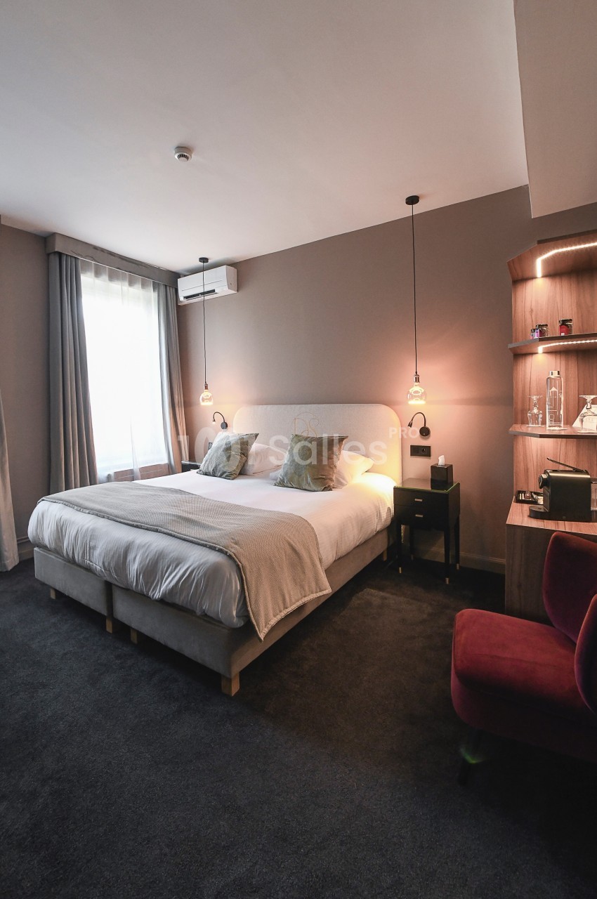 Chambre d'hôtel moderne avec un grand lit double, éclairage suspendu, fauteuil rouge et étagères en bois.
