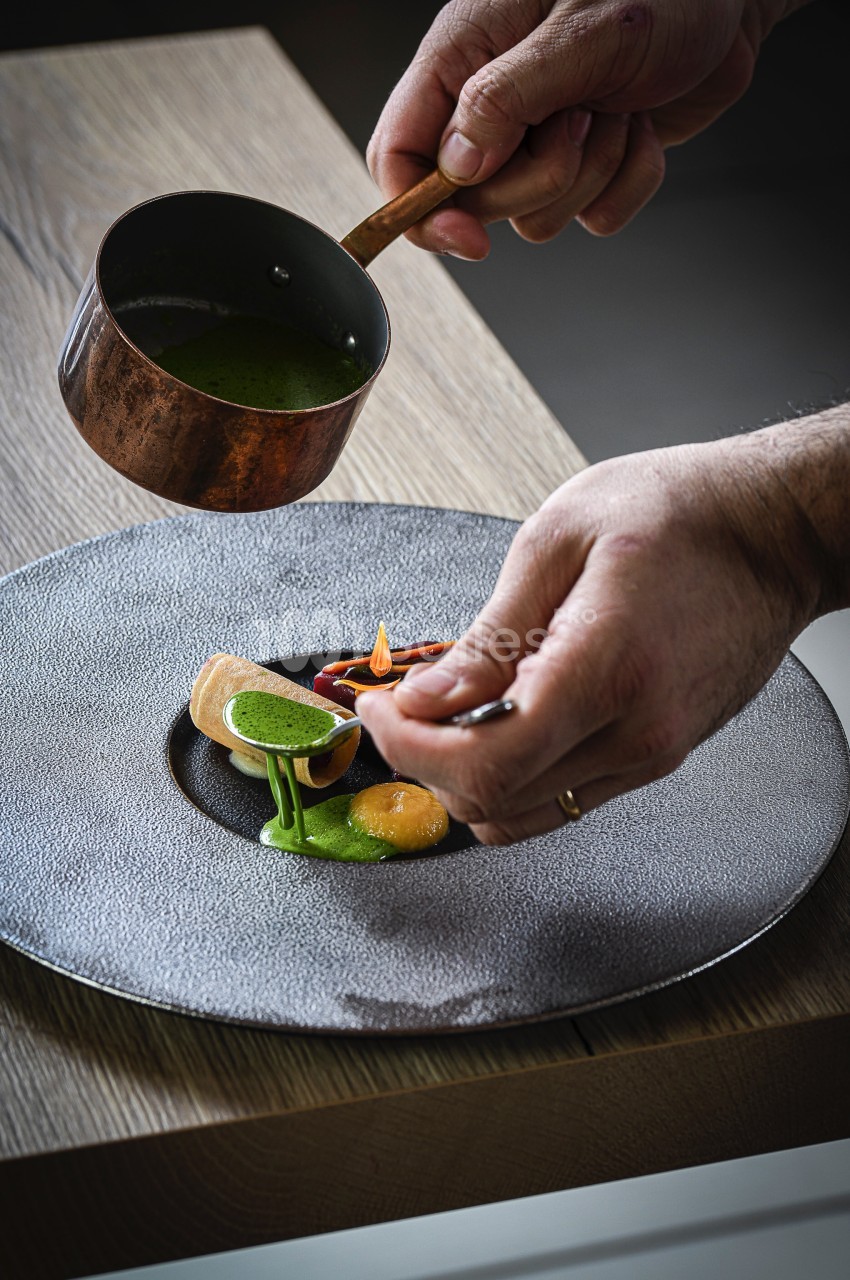 Un chef verse une sauce verte sur un plat raffiné présenté sur une assiette grise texturée.