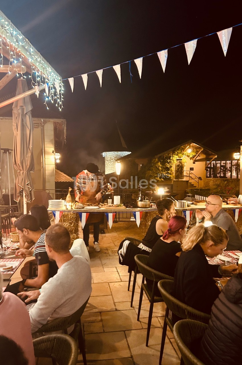 Terrasse animée d'un restaurant en soirée, avec des convives attablés et un cuisinier préparant des plats en extérieur.