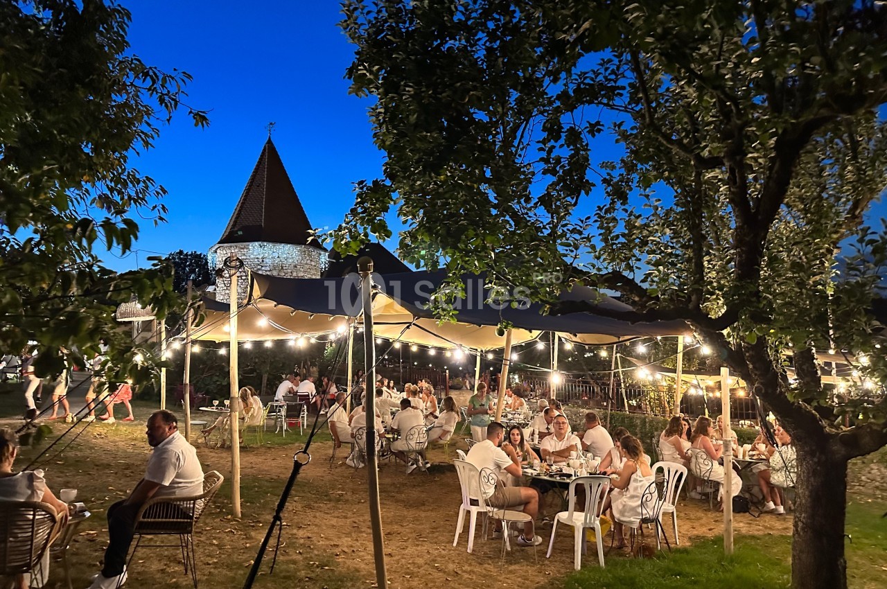 Personnes dînant en plein air sous une tente éclairée, entourées d'arbres, avec une église en arrière-plan au crépuscule.