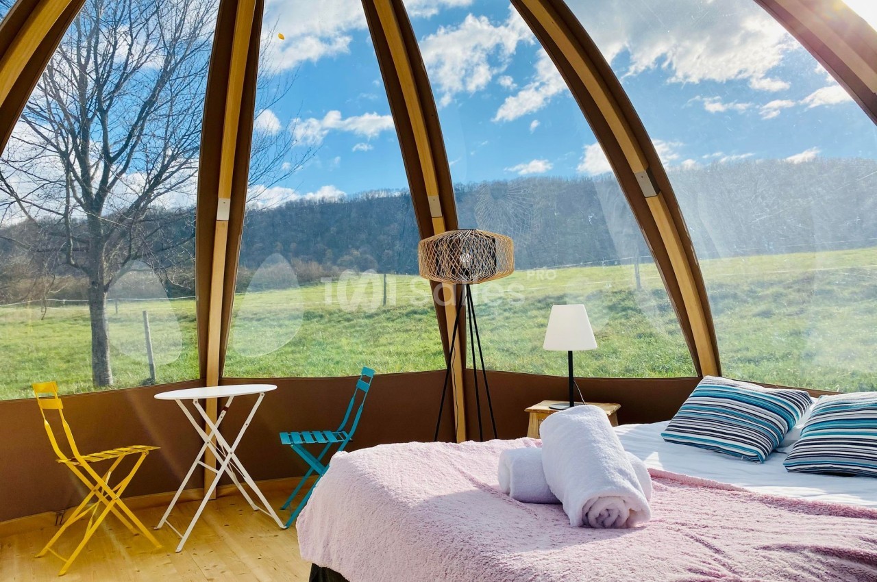 Intérieur d'une bulle avec lit, table et chaises, offrant une vue panoramique sur un paysage champêtre ensoleillé.