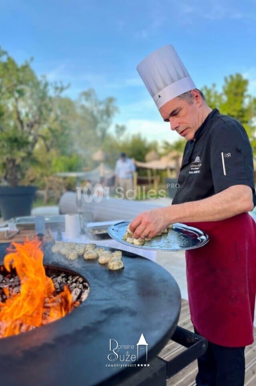 Un chef en tenue professionnelle cuisine des aliments sur une plancha en plein air près d'une piscine.