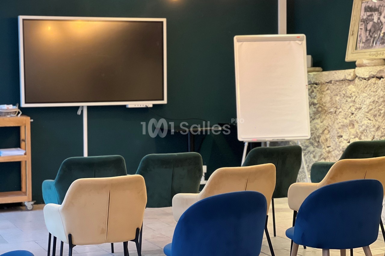 Salle de réunion avec chaises colorées, tableau blanc, écran et mur en pierre apparente.