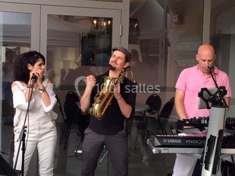 Trois musiciens jouent en extérieur : une chanteuse, un saxophoniste et un claviériste.