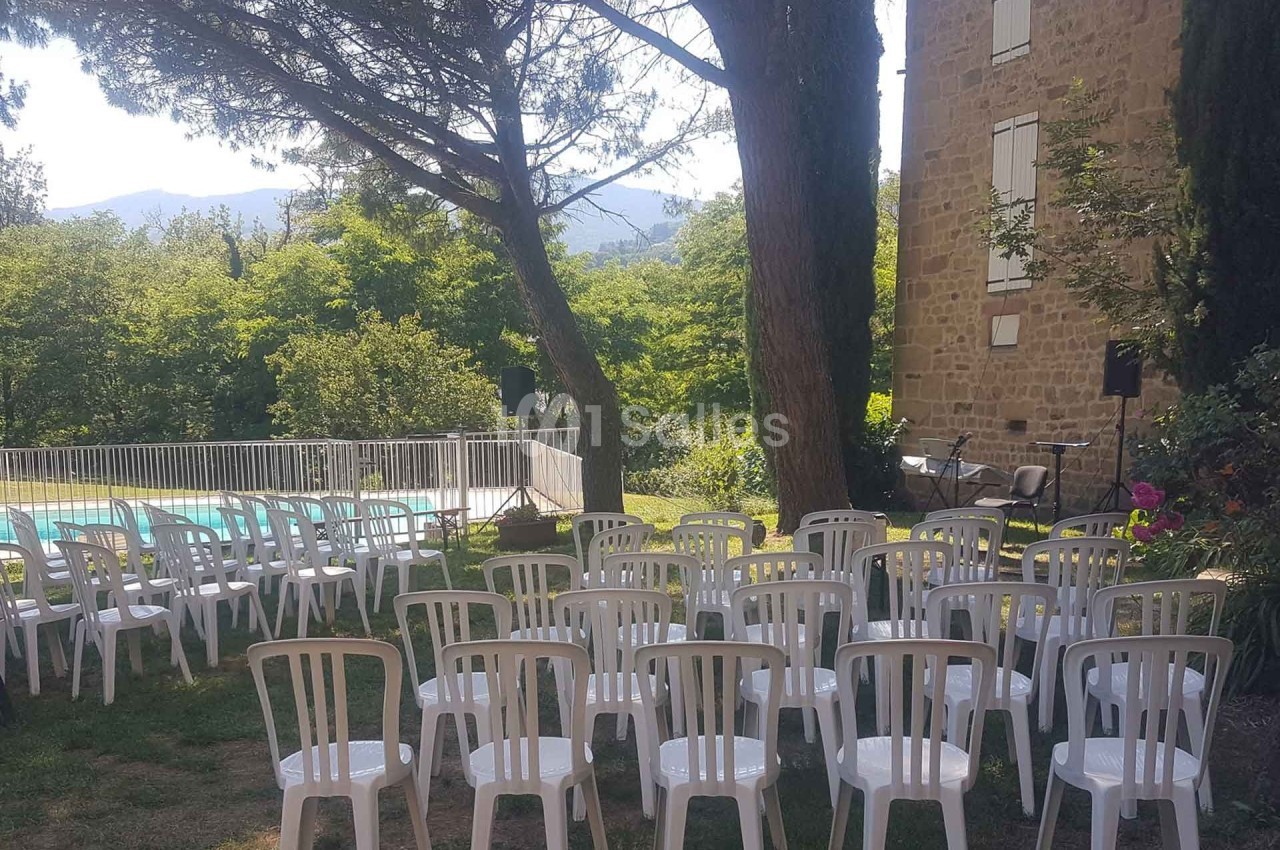 Chaises blanches disposées en extérieur près d'une piscine clôturée, entourées d'arbres et d'un bâtiment en pierre.