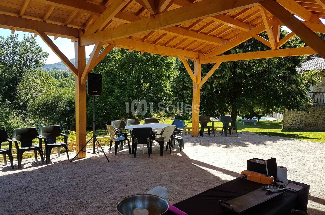 Structure en bois avec tables et chaises disposées sur un sol en gravier, entourée de verdure et d'arbres.