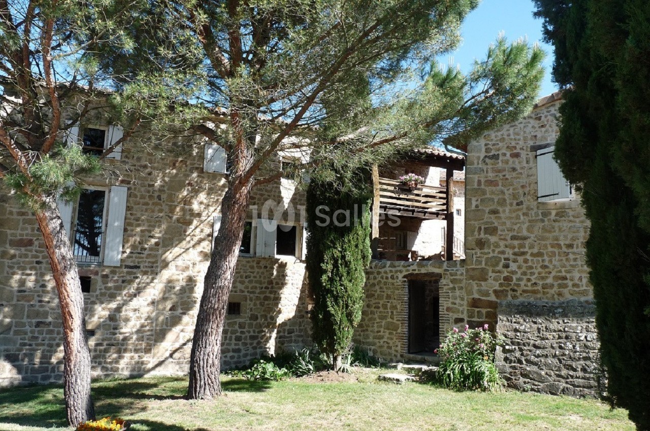 Façade en pierre d'une maison entourée d'arbres et d'un jardin verdoyant sous un ciel bleu.