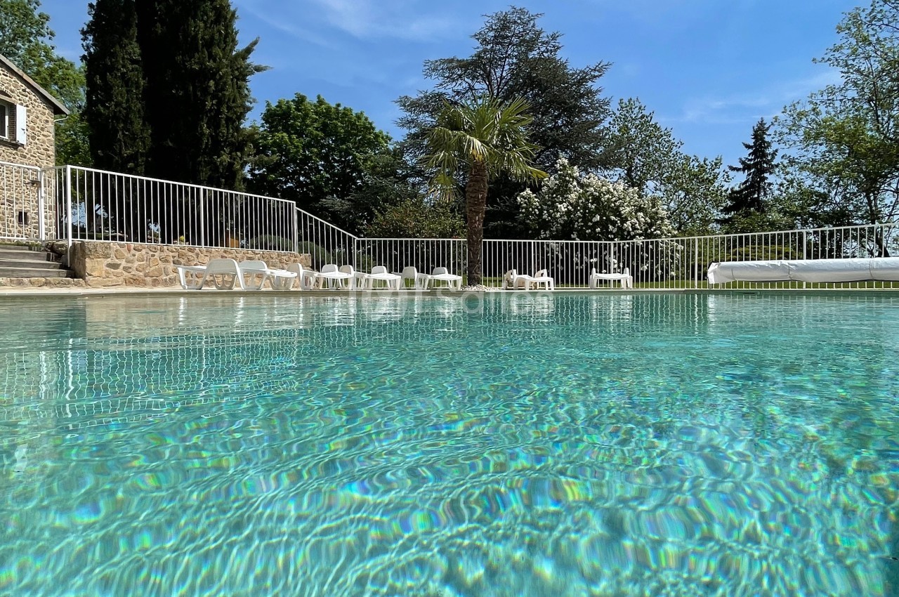 Piscine extérieure entourée de chaises longues blanches, bordée par une clôture et des arbres sous un ciel bleu.