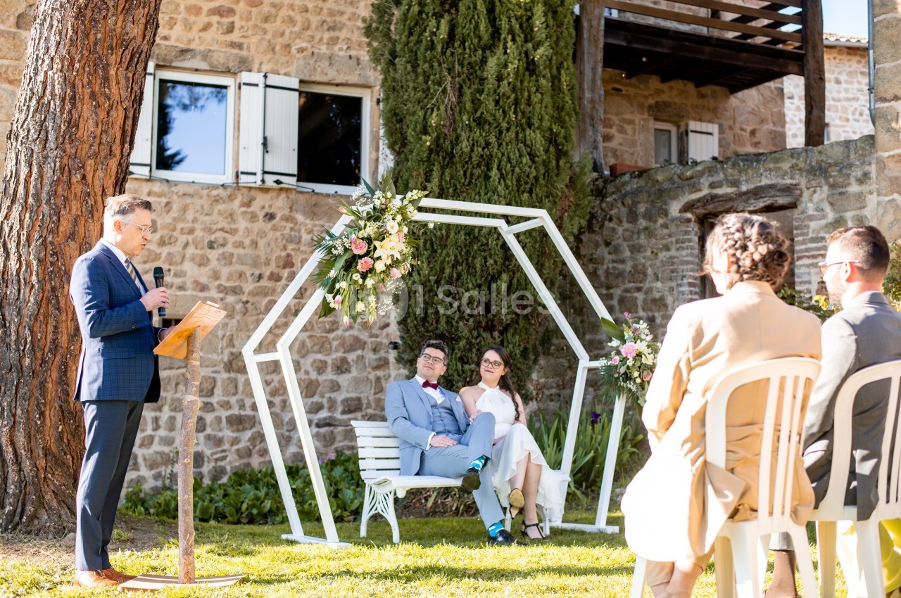 Un couple assis sous une arche décorée, écoutant un discours lors d'une cérémonie en extérieur près d'un bâtiment en pierre.