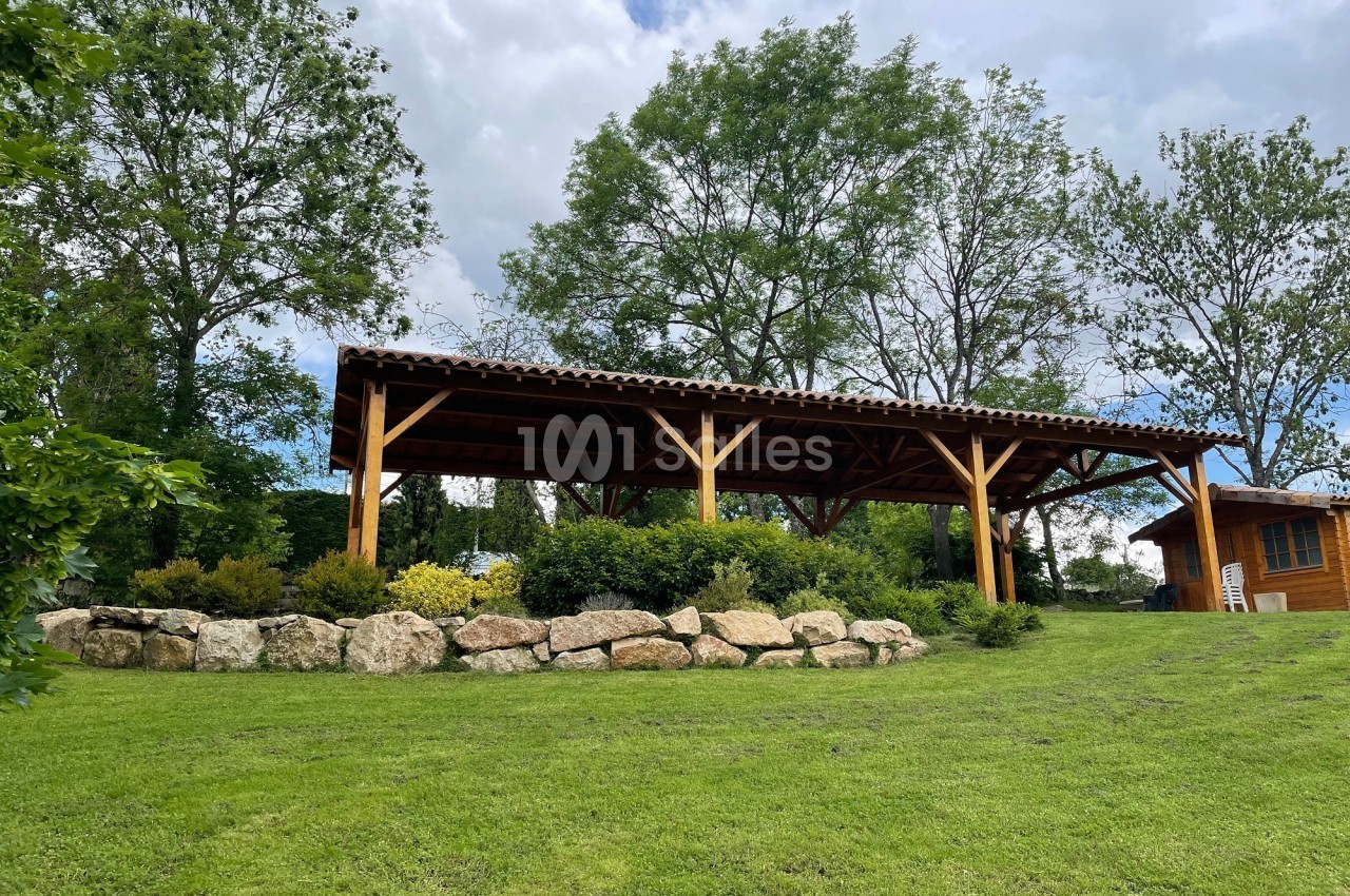 Abri en bois avec toit en tuiles, entouré de végétation et de rochers, situé sur une pelouse verdoyante.