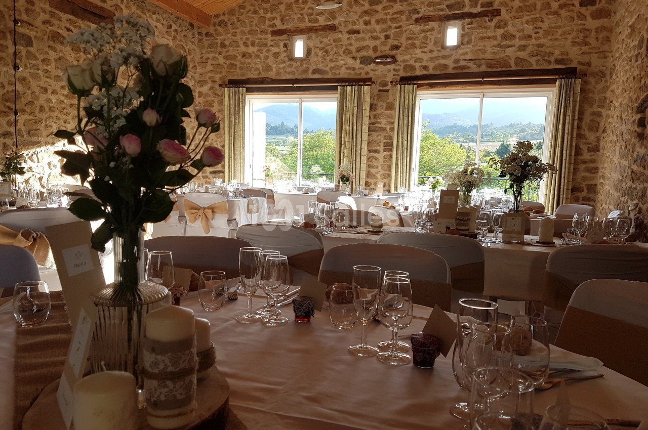 Salle de réception décorée pour un mariage, avec tables dressées, fleurs et vue sur un paysage extérieur verdoyant.