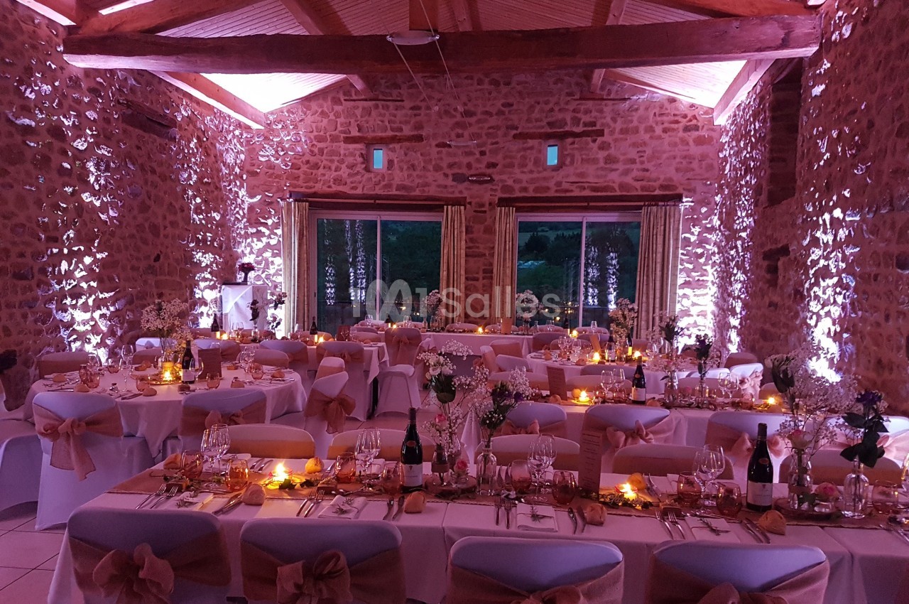 Salle en pierre décorée pour un mariage, avec tables dressées, nappes blanches, rubans beige et éclairage tamisé.