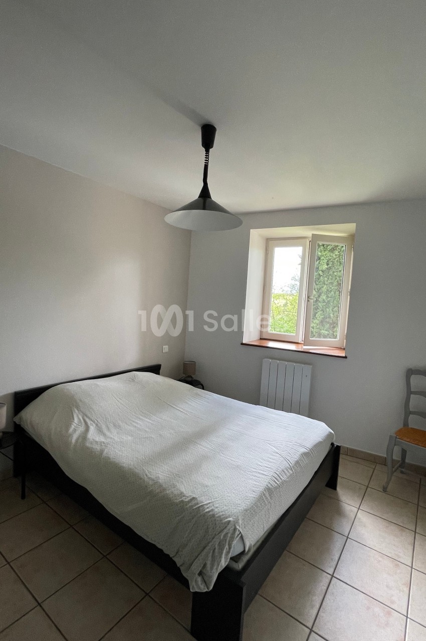 Chambre lumineuse avec un lit double, une suspension noire, une chaise grise et une fenêtre donnant sur un jardin.