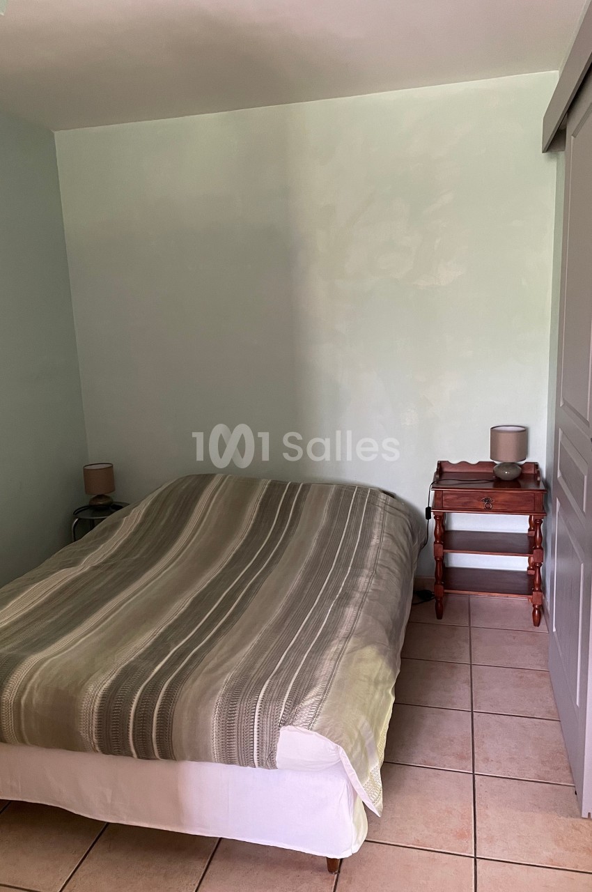 Chambre avec un lit double, draps rayés verts, table de chevet en bois, lampes et sol carrelé beige.