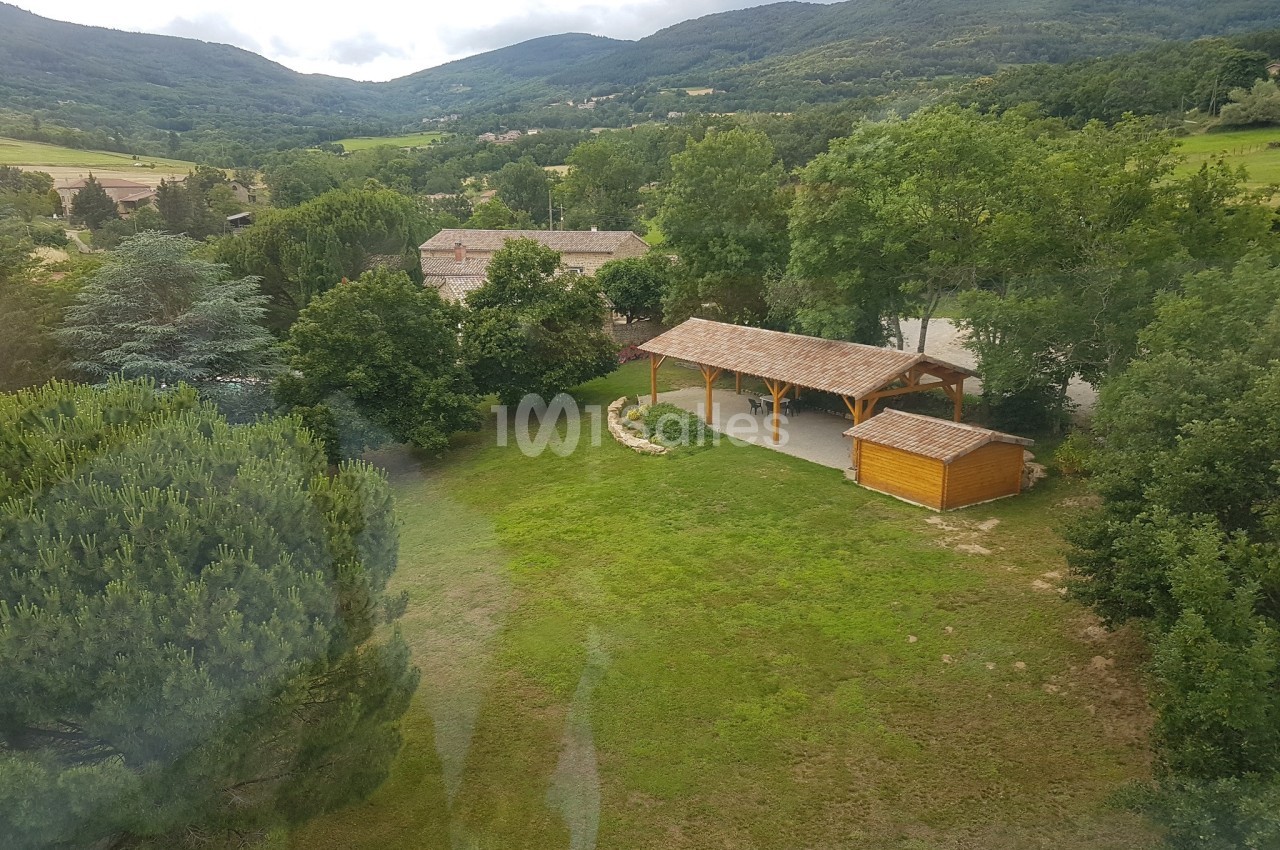 Vue aérienne d'un espace verdoyant avec un abri en bois, des arbres et des collines en arrière-plan.