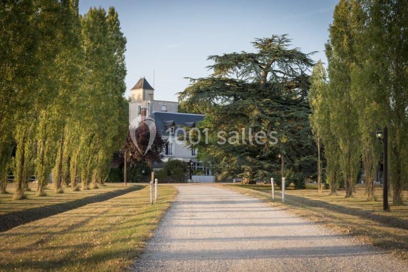 Allée bordée d'arbres menant à une maison en pierre avec une tour, entourée de verdure.