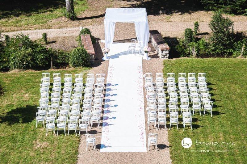 Allée centrale bordée de chaises blanches menant à une arche de mariage décorée de tissu blanc, en extérieur.