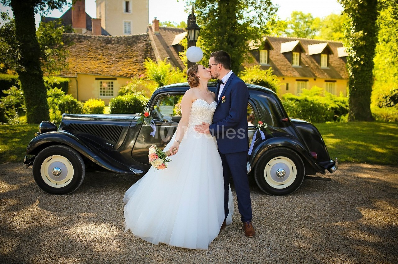 Un couple en tenue de mariage s'embrasse devant une voiture ancienne, dans un cadre verdoyant et ensoleillé.