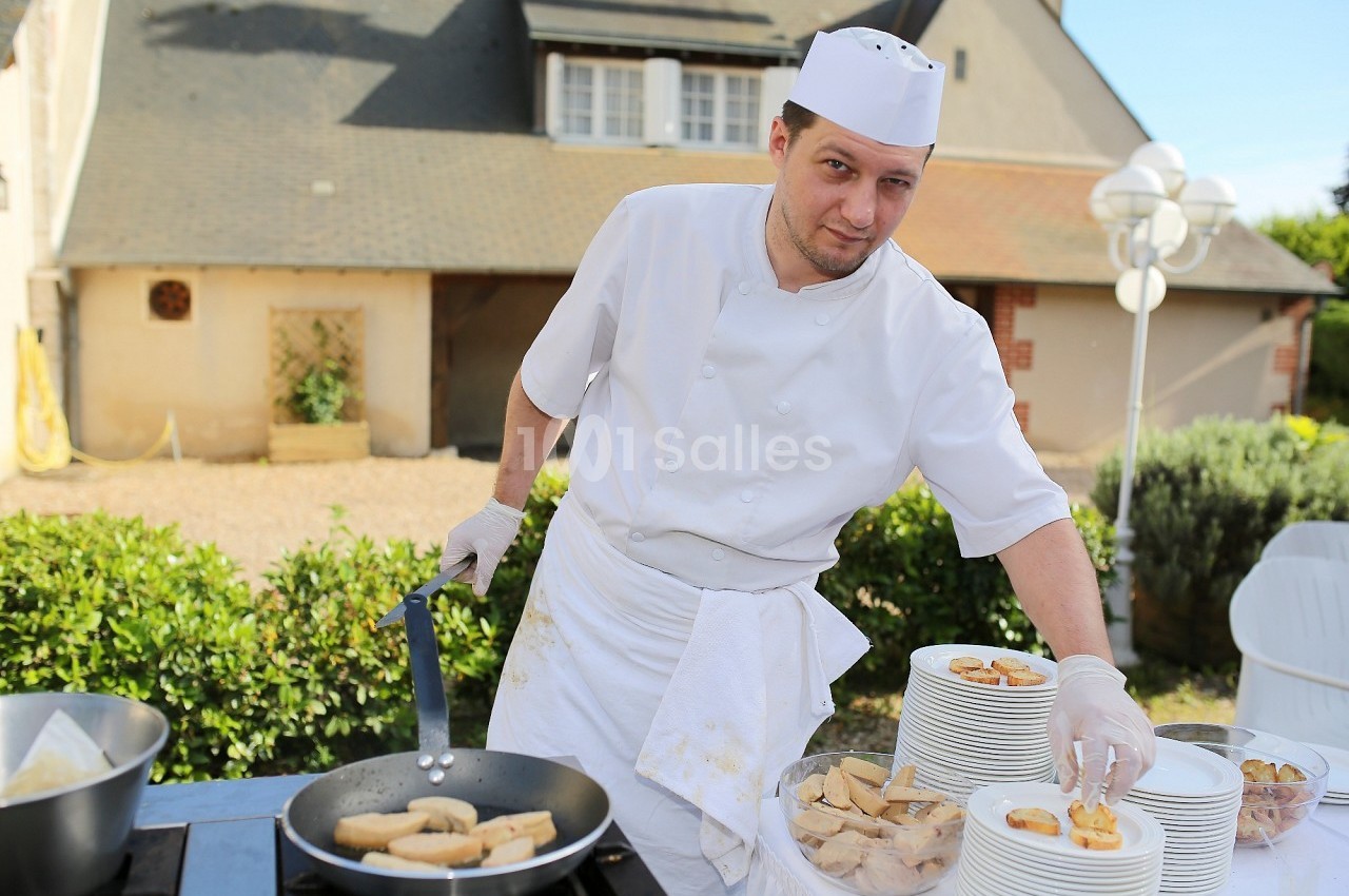 Un cuisinier en tenue blanche prépare des beignets dans une poêle en extérieur, près de piles d'assiettes.