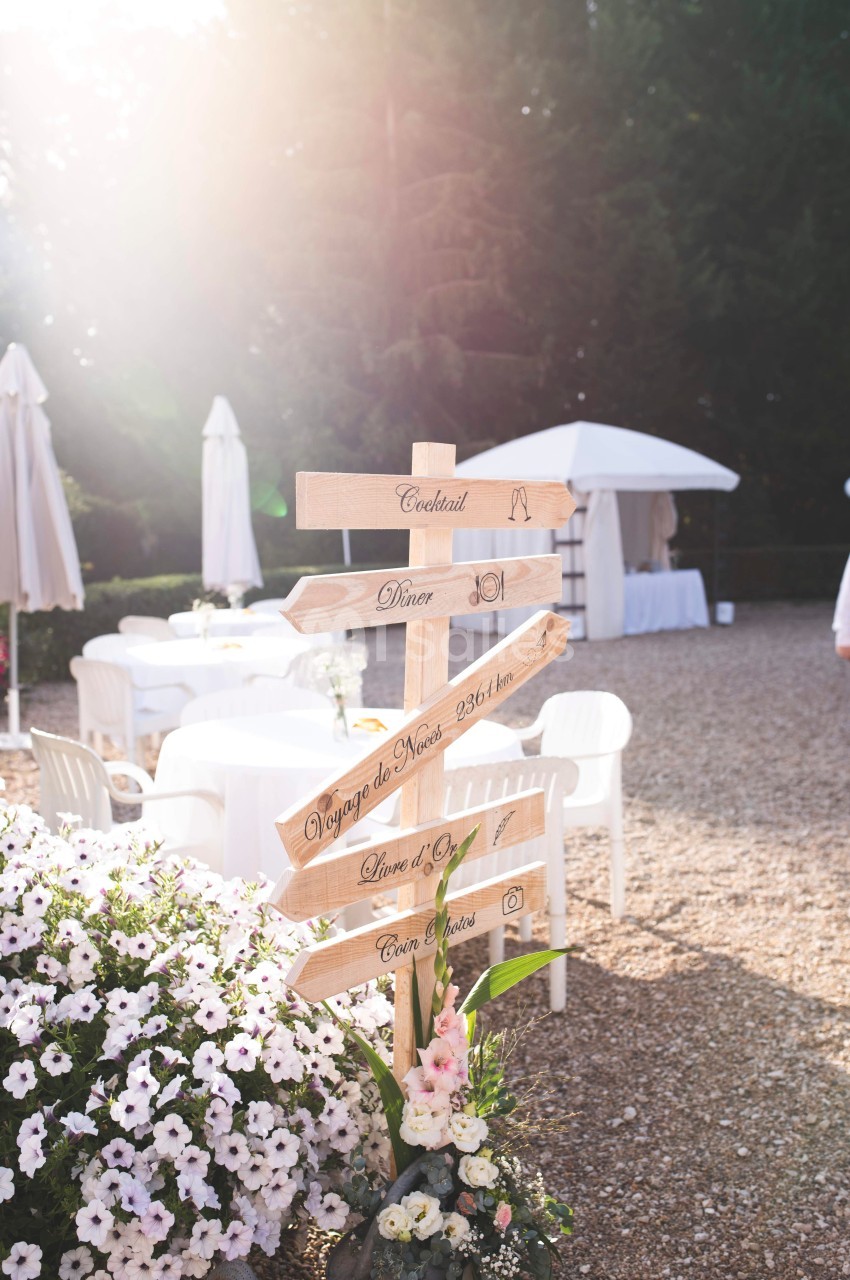 Panneau en bois indiquant différentes directions lors d'un événement en extérieur, entouré de fleurs et de mobilier blanc.