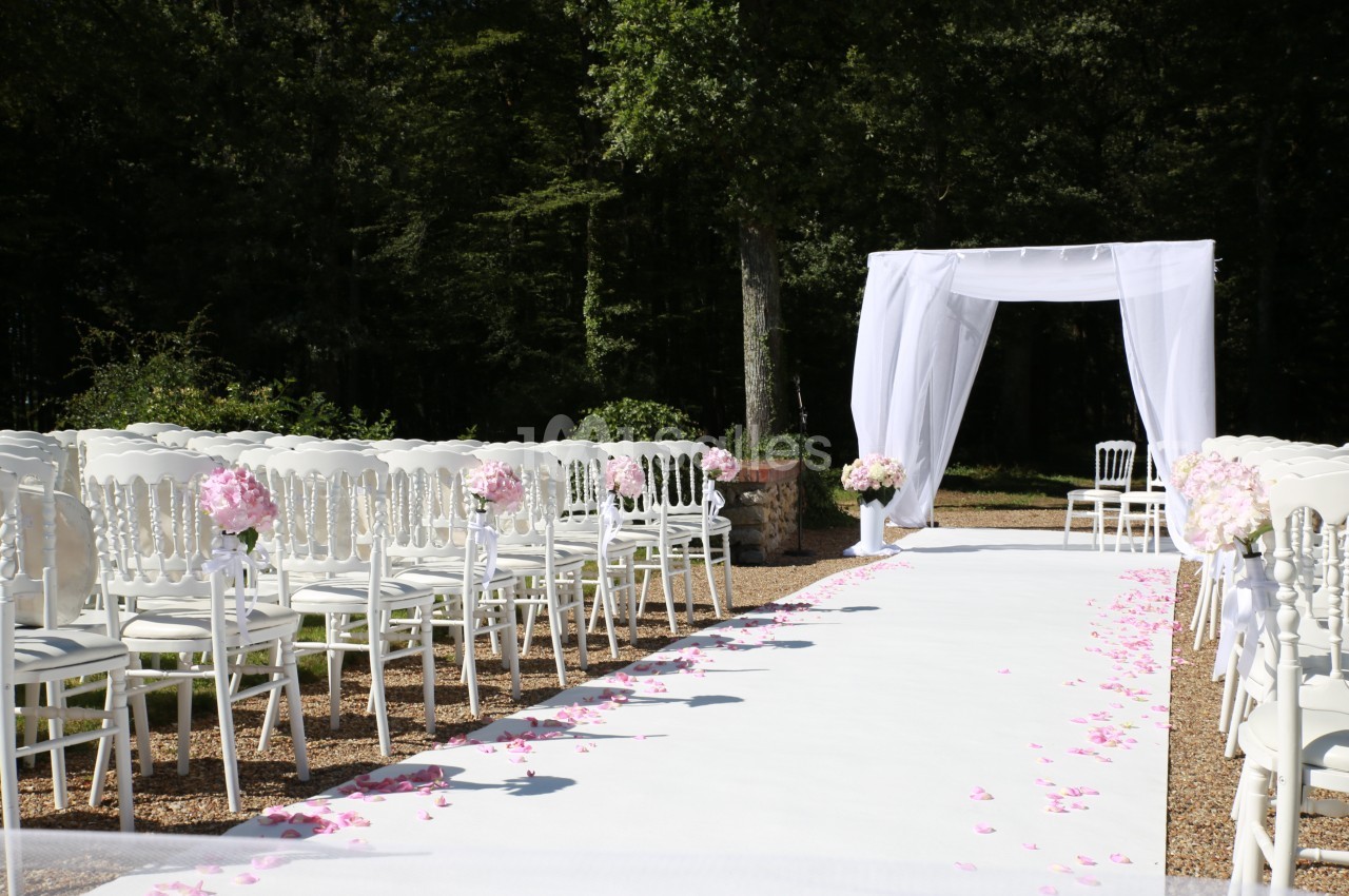 Allée centrale décorée de pétales roses, entourée de chaises blanches, menant à une arche blanche en extérieur.