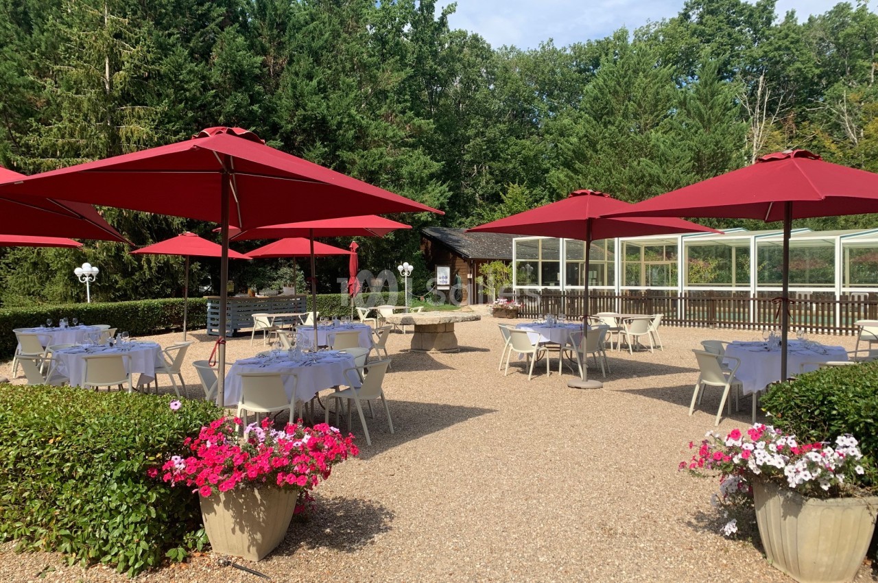 Terrasse extérieure avec tables et chaises blanches sous des parasols rouges, entourée de verdure et de fleurs.