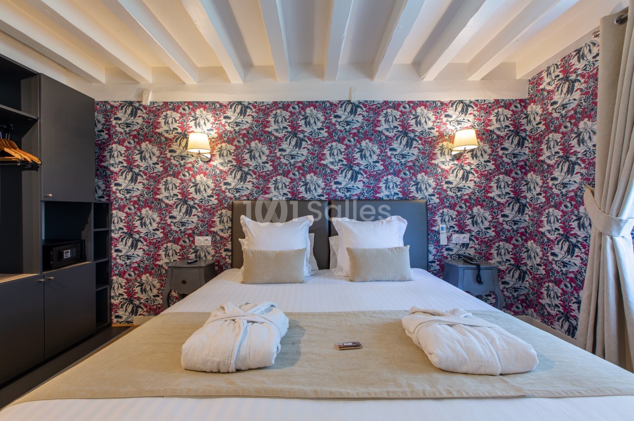 Lit double avec draps blancs, coussins beiges, peignoirs pliés, mur fleuri coloré et éclairage mural doux.