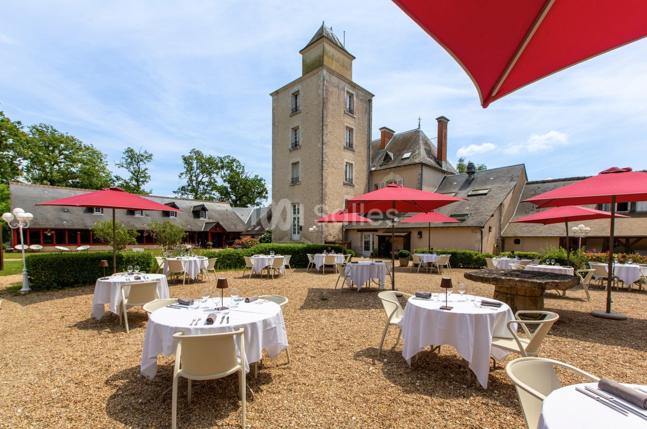 Terrasse d'un restaurant avec tables dressées sous des parasols rouges, située dans une cour devant un bâtiment ancien.