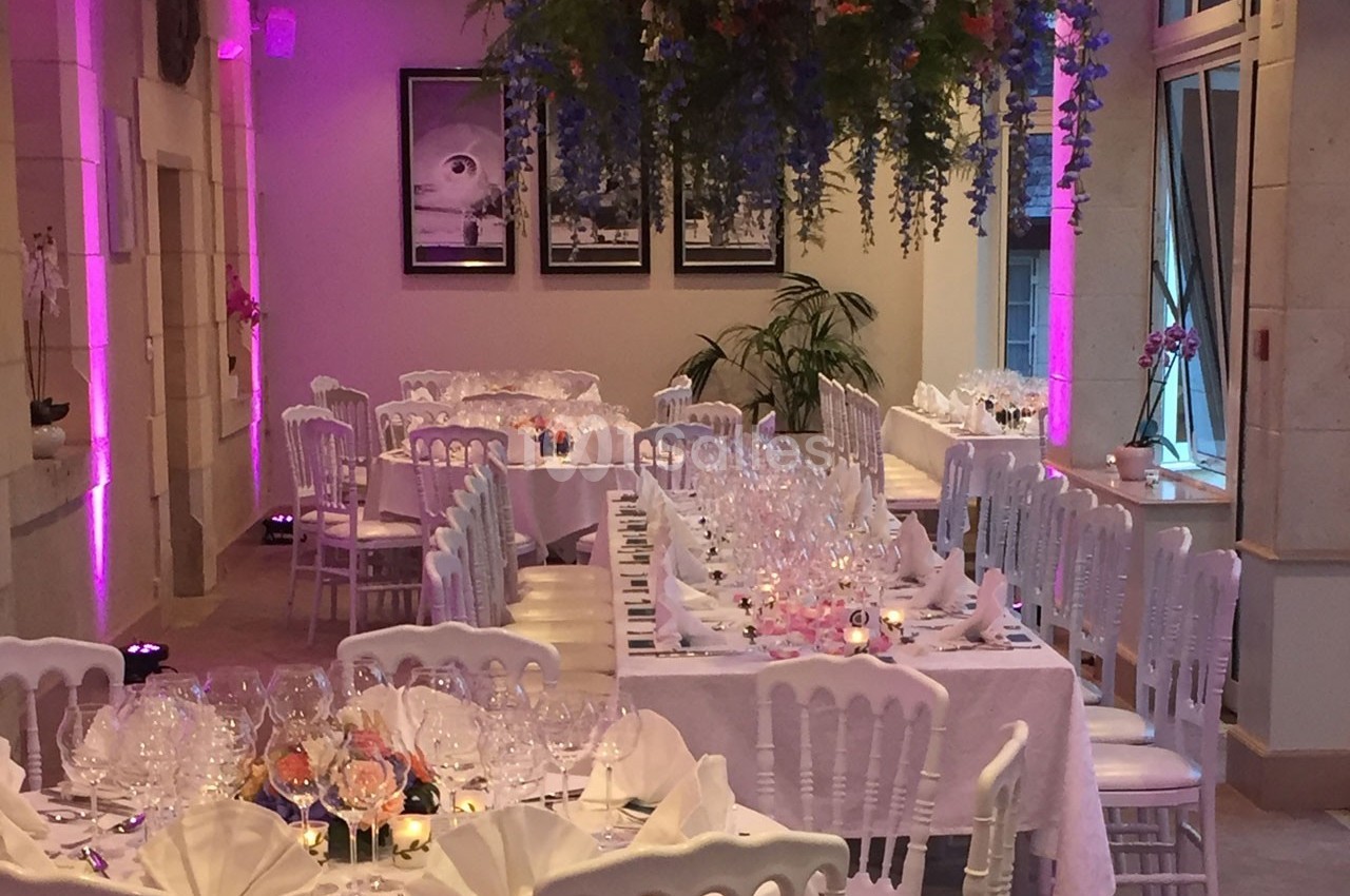 Salle de réception décorée avec des tables dressées, des chaises blanches et des fleurs suspendues au plafond.