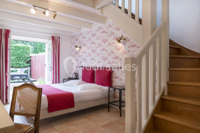 Chambre lumineuse avec lit double, escalier en bois, murs décorés de papier peint floral et accès à une terrasse extérieure.