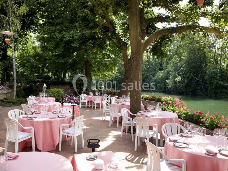Terrasse d'un restaurant avec des tables dressées sous des arbres, près d'une rivière entourée de verdure.