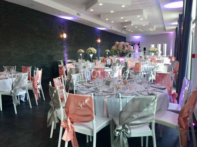 Salle de réception élégamment décorée avec des tables rondes, des chaises ornées de nœuds roses et argentés, et des centres…