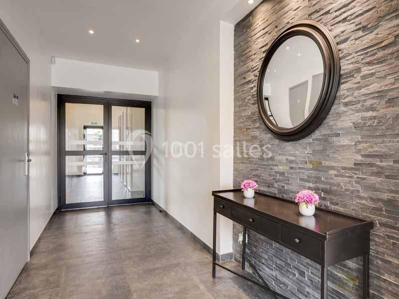 Entrée moderne avec mur en pierre, miroir rond, table noire décorée de fleurs roses et porte vitrée au fond.