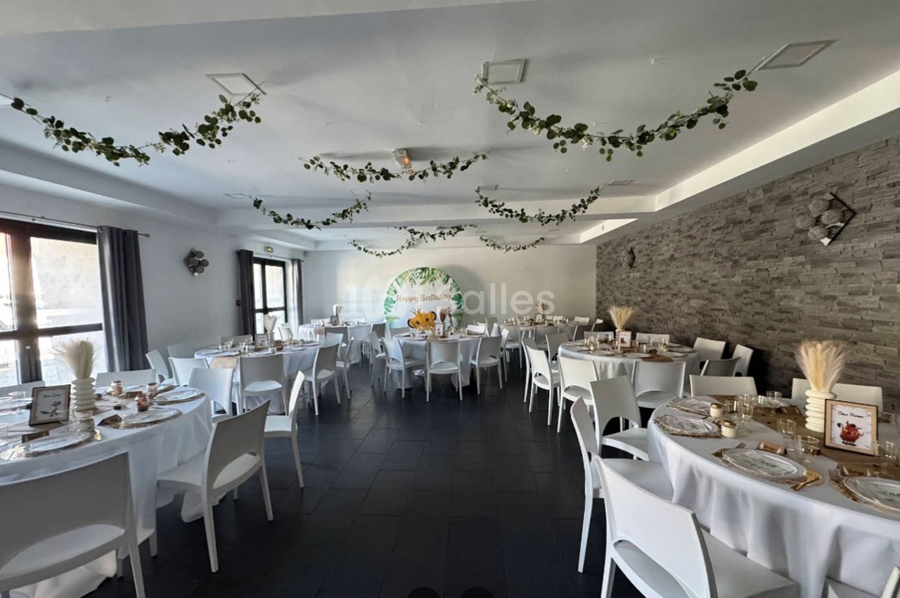 Salle de réception décorée avec des tables rondes, nappes blanches, guirlandes végétales et un mur en pierre.