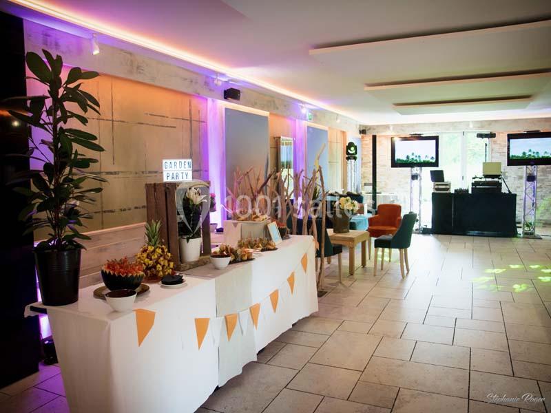 Salle décorée pour un événement avec table de buffet, plantes, éclairage coloré et espace DJ en arrière-plan.
