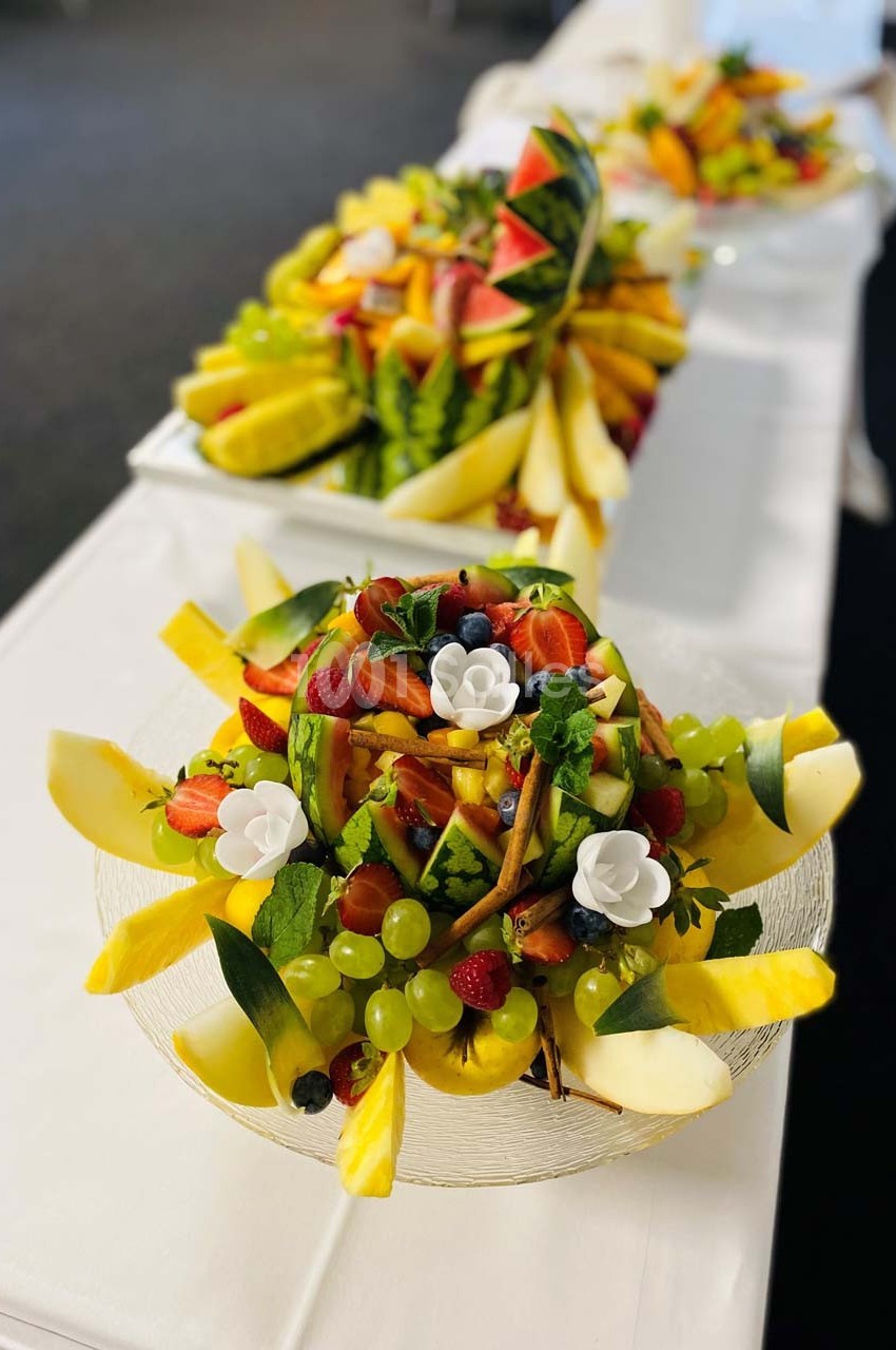 Plateaux de fruits frais joliment disposés avec des morceaux de pastèque, ananas, raisins, fraises et décorations florales.