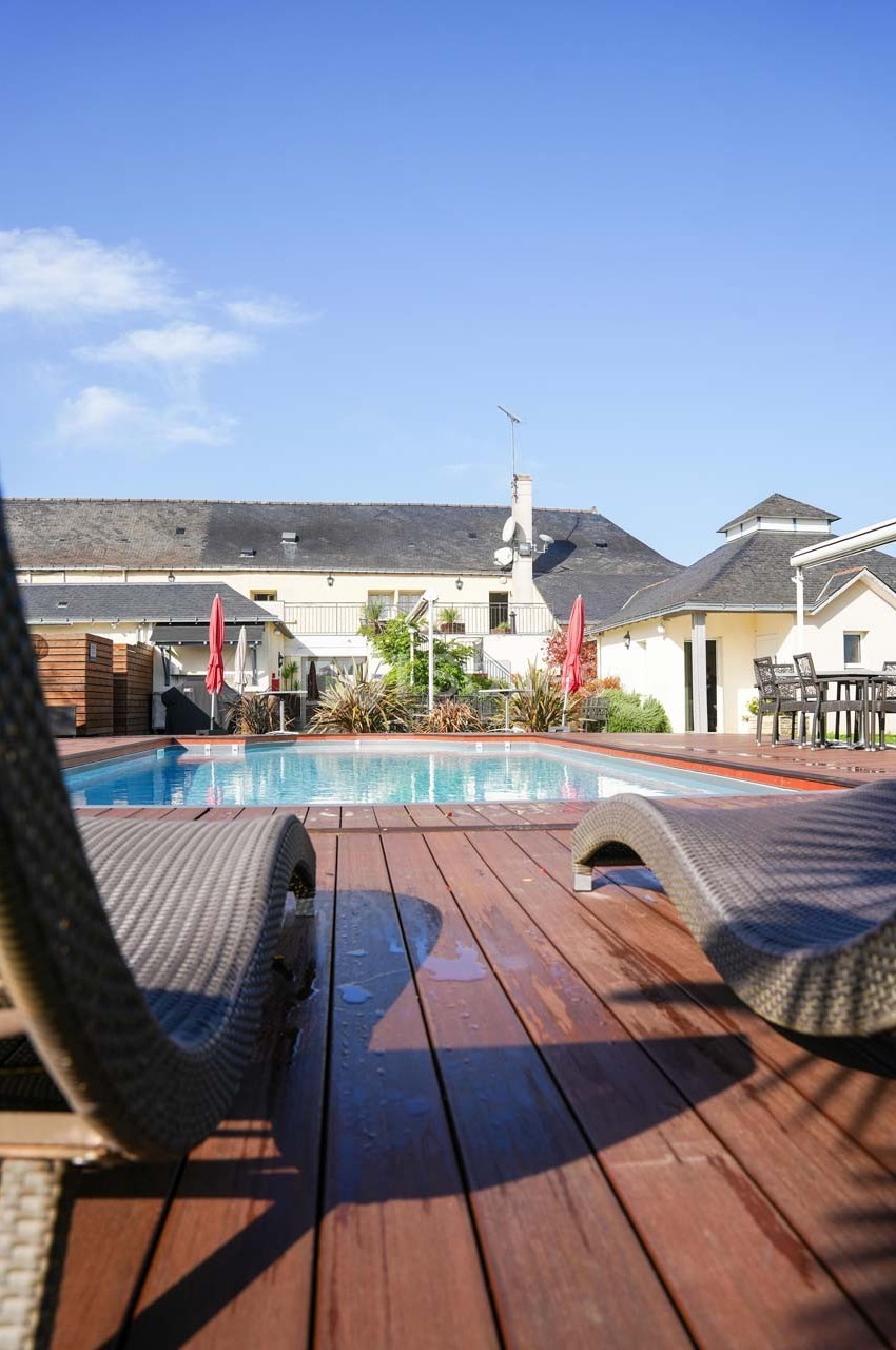 Piscine extérieure entourée de chaises longues et parasols, avec des bâtiments en arrière-plan sous un ciel dégagé.