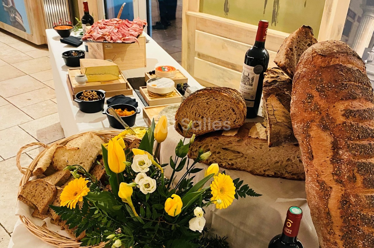 Table garnie de pains variés, charcuteries, fromages, fleurs jaunes et blanches, accompagnée de bouteilles de vin rouge.
