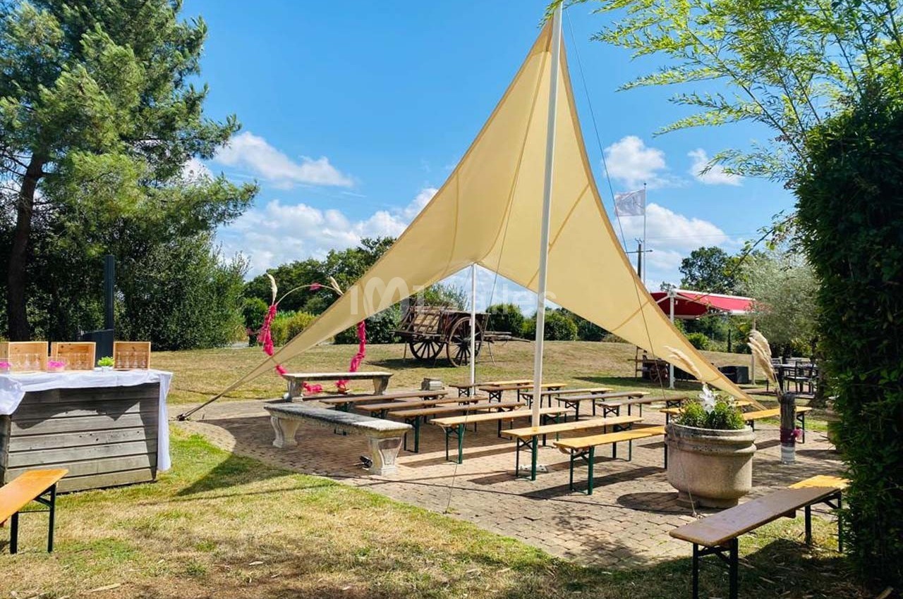 Tables et bancs en bois disposés sous une voile d'ombrage dans un espace extérieur arboré et ensoleillé.