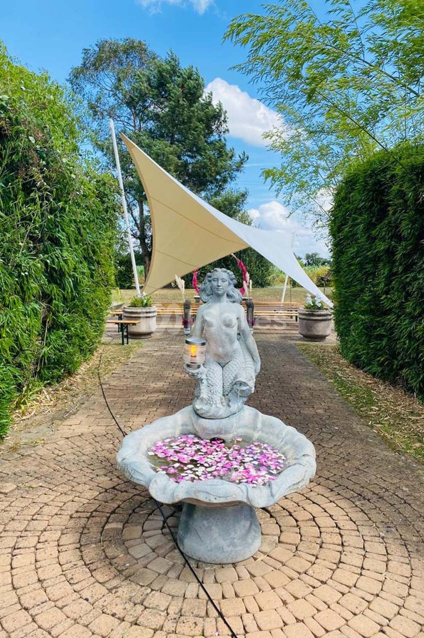 Statue de sirène tenant une lampe, placée dans une fontaine ornée de fleurs, sur une allée pavée entourée de verdure.