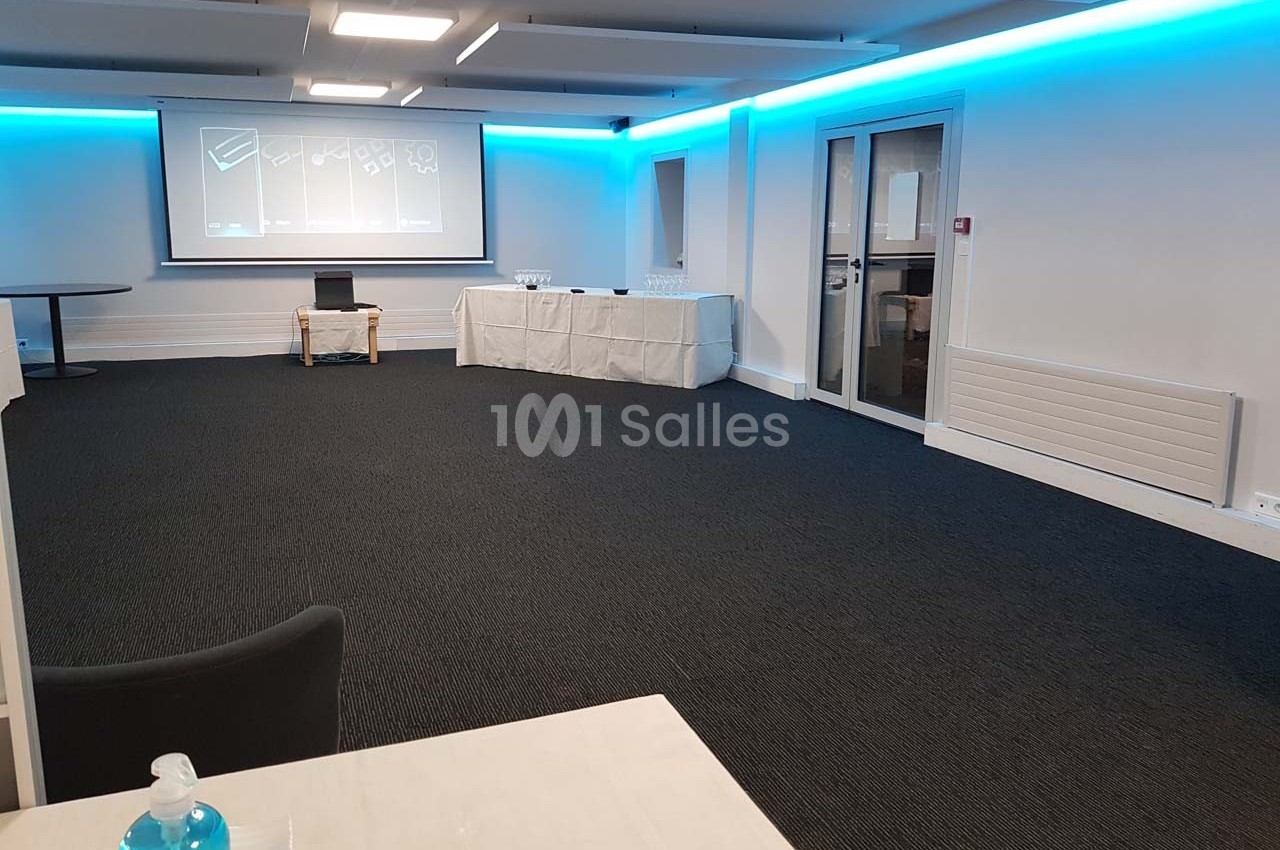 Salle de réunion vide avec un projecteur allumé, des tables couvertes de nappes blanches et des chaises disposées autour.