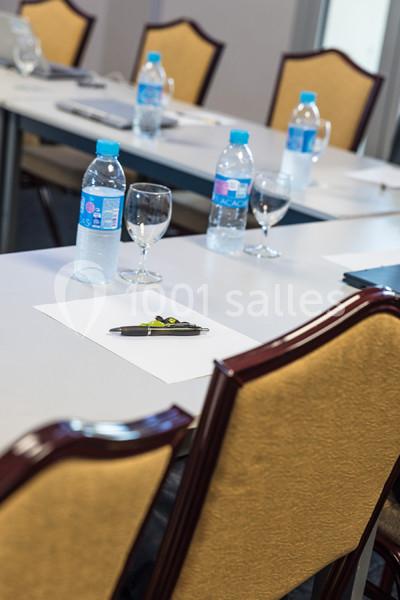 Salle de réunion avec des chaises, des bouteilles d'eau, des verres et des stylos posés sur des tables blanches.