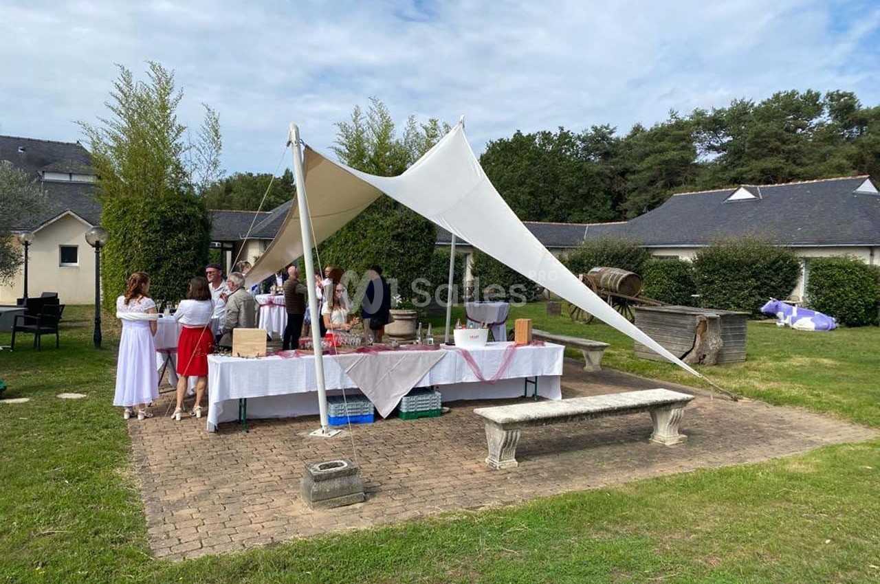 Des personnes rassemblées autour d'une table dressée sous une toile blanche dans un jardin avec des bâtiments en arrière…