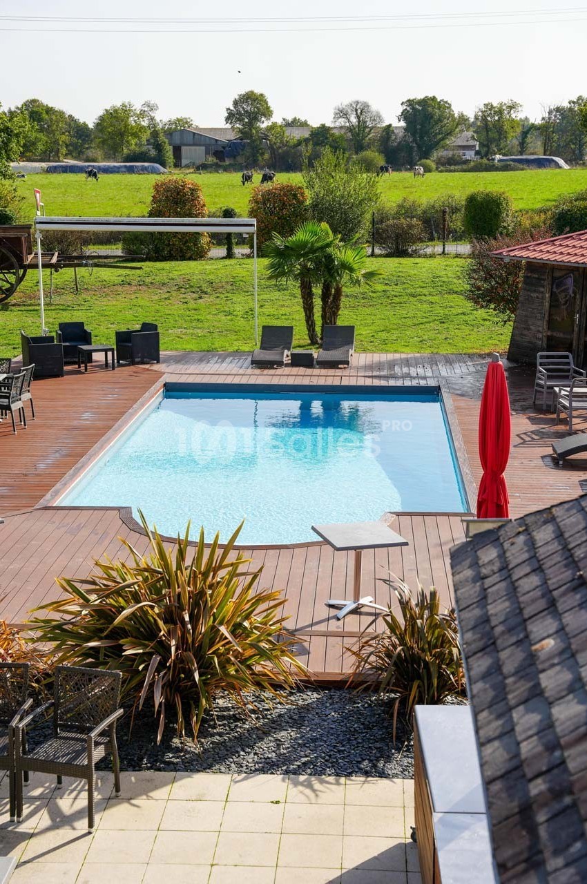 Piscine extérieure entourée de terrasses en bois, chaises longues et végétation, avec un champ en arrière-plan.