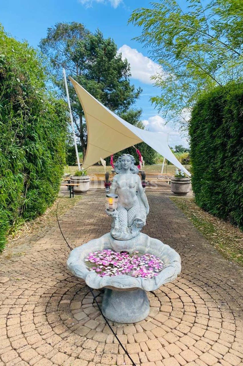 Statue de sirène tenant une lampe, placée devant une fontaine remplie de fleurs, dans une allée bordée de verdure.