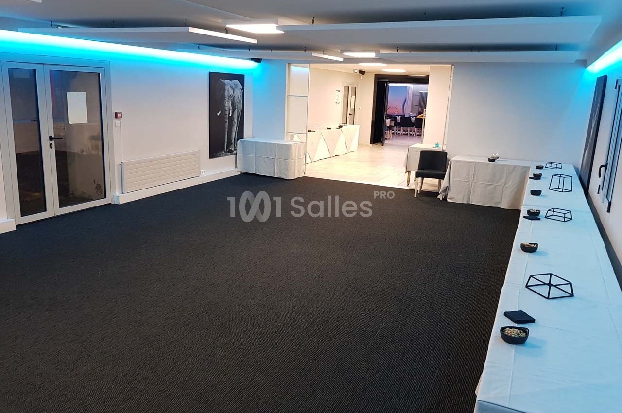 Salle vide avec moquette sombre, tables alignées le long des murs, éclairage bleu et porte vitrée sur la gauche.