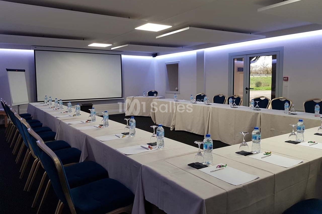 Salle de réunion avec tables disposées en U, chaises alignées, écran de projection, paperboard et bouteilles d'eau.