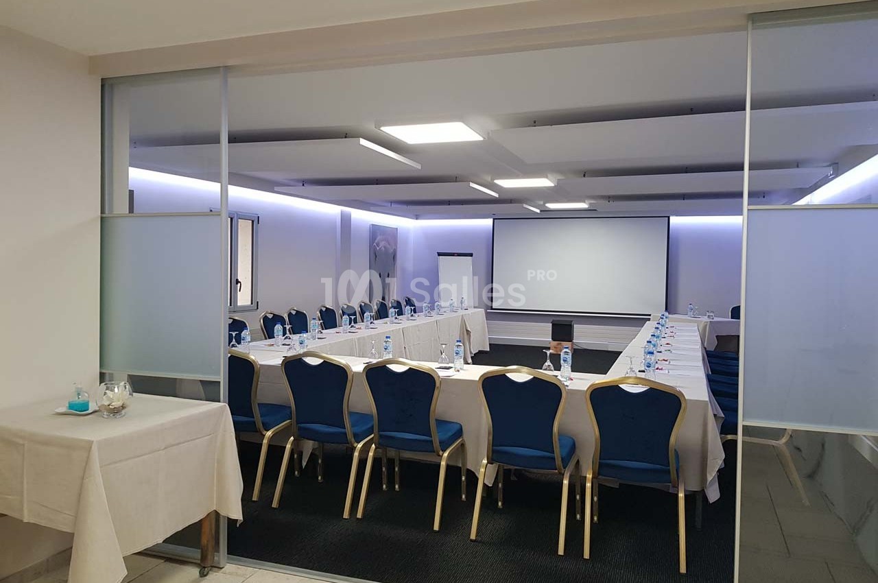 Salle de réunion équipée de tables en U, chaises bleues, écran de projection et éclairage moderne.