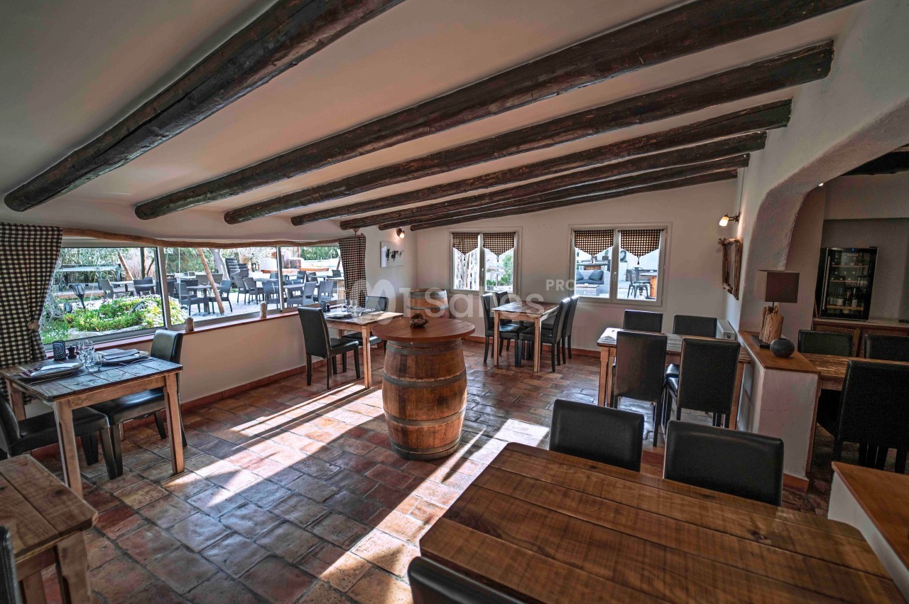Salle de restaurant lumineuse avec poutres apparentes, tables en bois et tonneau central, vue sur terrasse extérieure.